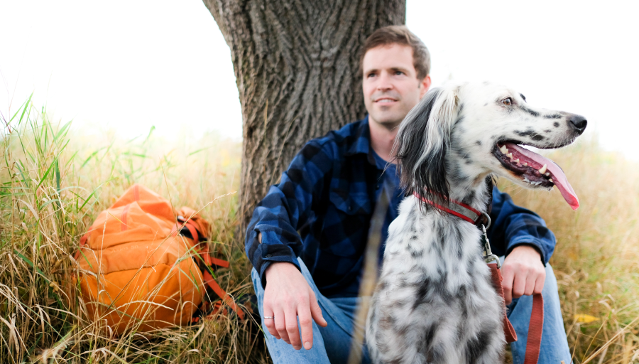 Uomo seduto di fronte a un albero con il suo zaino arancione su un prato e il suo cane al guinzaglio di fronte a lui
